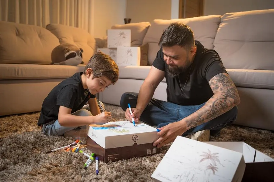 Pai e filho desenhando em caixa de doces no tapete da sala, campanha Sodiê Doces